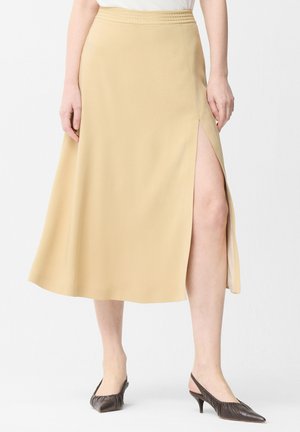 Femme portant une jupe midi beige avec une fente latérale et des escarpins slingback à bout pointu marron foncé, debout devant un fond blanc uni.