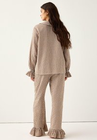 Kvinde med langt brunt hår iført brun-og-hvid ternede pyjamas med flæser ved ærmerne, som står barfodet foran en enkel hvid baggrund.