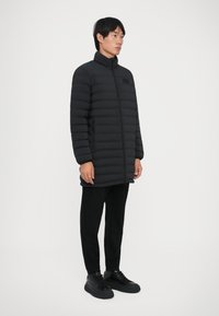 Giovane uomo in piedi, che indossa un lungo giubbotto nero trapuntato, pantaloni neri e sneakers nere, che guarda di lato su uno sfondo neutro.