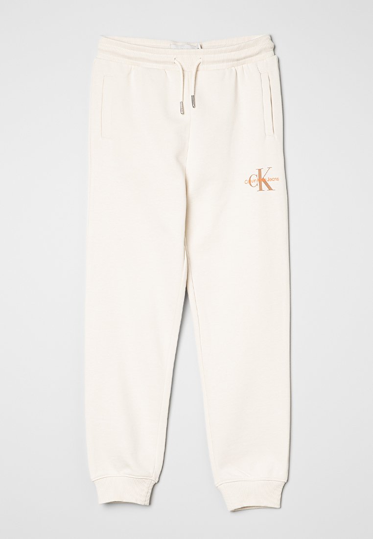 Pantalons de survêtement couleur crème avec taille élastique, bretelles et poches latérales. Présente le logo "CK" en cuivre sur la cuisse gauche. Fabriqué en tissu doux.