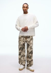 Muž stojí a má na sobě přehnaně velkou bílou košili s dlouhým rukávem, široké kalhoty s abstraktním vzorem, bílé tenisky a tenký náhrdelník.