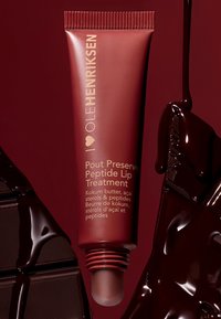 Tubo marrón con una tapa estriada, con texto dorado. Tratamiento labial etiquetado como "Pout Preserve Peptide." Fondo brillante de chocolate oscuro.