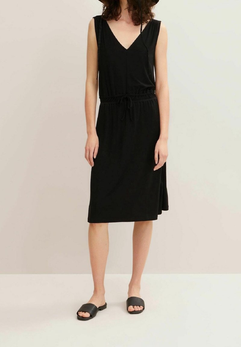 Femme portant une robe midi noire sans manches avec un col en V et une taille à cordon, associée à des sandales noires à enfiler, debout devant un fond neutre.