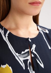 Námořnické modré šaty s kulatým výstřihem, se zapínáním na knoflíky a květinovým vzorem v bílé a žluté barvě, vyrobené z hladké tkaniny.