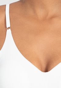 Witte bralette met dunne bandjes, V-hals ontwerp, gladde textuur en een klein zilveren ringdetail op elk bandje. Huidskleurige achtergrond.