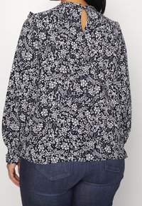 Blusa floreale blu navy con maniche lunghe a sbuffo, caratterizzata da motivi botanici bianchi e un'apertura a chiave sul retro. Tessuto morbido, vestibilità comoda.