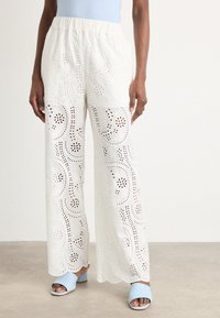 Pantaloni ampi bianchi ricamati presentano motivi floreali a taglio, una vita elastica e orli scallop. Abbinati a tacchi azzurri chiari.
