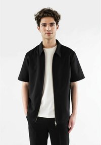 Svart kortärmad zip-up skjorta med spetsig krage över en vit t-shirt, mjukt tyg och minimalistisk design.