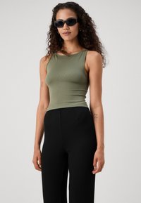 Olivově zelený žebrovaný top na ramínka s kulatým výstřihem, doplněný černými žebrovanými širokými kalhotami. Modelka má na sobě černé obdélníkové sluneční brýle.