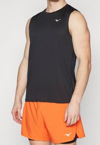 Černý bezrukávový sportovní top s hladkým povrchem, s bílým logem na hrudi, v kombinaci s oranžovými šortkami.