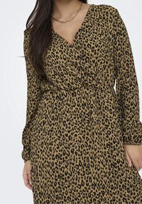 Kleid mit Leopardenmuster, V-Ausschnitt, langen Ärmeln und tailliertem Schnitt. Der Stoff hat eine weiche Textur mit einem sandfarbenen Hintergrund und schwarzen Flecken.