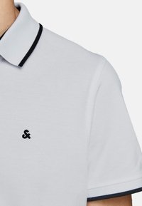 Witte polo met een gestructureerde stof, voorzien van een zwarte kraag en mouwboorden, en een klein zwart logo op de linkerkant van de borst.