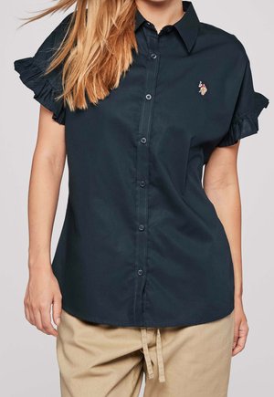 Femme portant une chemise bleu marine à manches courtes avec des manches volantées et un pantalon beige, les cheveux tombant sur l'épaule gauche.