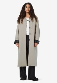 Vrouw met lange krullende haren in een beige lange jas met zwarte manchetten, witte top, zwart-witte broek en zwarte schoenen die staat.