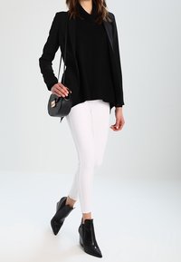Schwarzer Blazer über einem schwarzen Oberteil, kombiniert mit weißen skinny Jeans und schwarzen Wedge-Stiefeletten. Das Model hält eine kleine runde schwarze Handtasche.