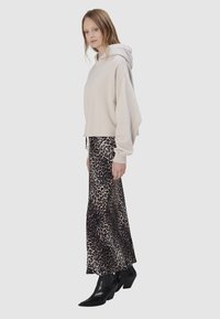 Vrouw in een beige oversized hoodie, een maxi-rok met luipaardprint en zwarte enkellaarsjes met hak, staand tegen een egale lichte achtergrond.