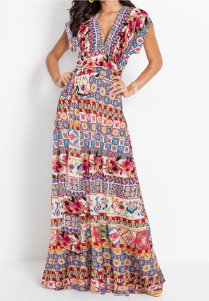 Femme portant une robe longue patchwork florale colorée avec des manches courtes volantées et un décolleté en V, mains sur les hanches sur fond blanc.