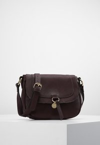 Tmavě hnědá kožená crossbody taška s nastavitelným popruhem, klopovým uzávěrem a malým kulatým zlatým přívěskem vpředu, vystavená na bílé podstavě.