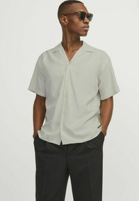 Light gray short-sleeve shirt with a notched collar and button front. Worn with black trousers, model also wears dark sunglasses.