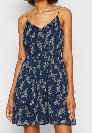 Žena v tmavě modrých šatech bez rukávů s tenkými ramínky a květinovým vzorem, ukazující tetování na obou předloktích.