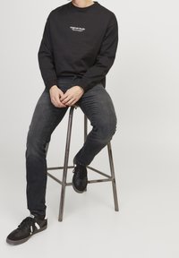 Camisa negra de manga larga con texto blanco, combinada con jeans oscuros ajustados y zapatillas negras, sentado en un taburete de metal contra un fondo blanco.