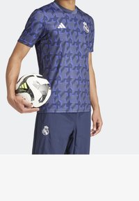 Muž držící černobílý fotbalový míč Adidas, oblečený v tmavě modrých šortkách a modrém tréninkovém tričku Real Madrid s vzorem.