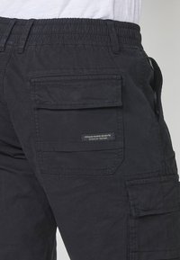 Pantalons cargo noirs avec une taille élastique, plusieurs poches latérales et une étiquette indiquant la marque et le numéro de stock. Matière en coton texturé.