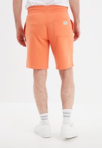 Shorts de algodón naranja con etiqueta en el bolsillo trasero. Presentan una cintura elástica y un ajuste relajado, que terminan justo por encima de la rodilla.