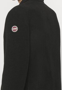 Veste en polaire noire avec un patch rond du logo Colmar sur l'épaule. Le design est simple avec une finition douce et texturée.