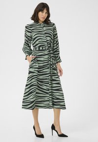 Zeleno oblečeno s črnimi zebra črtami, s dolgimi rokavi in gumbi. Ima zavezan pas, žepe in sproščen kroj. Gladek občutek tkanine.