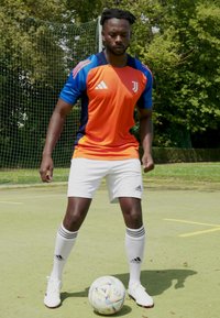 Camisa de futebol laranja e azul da Adidas com logo, calções brancos e meias às riscas, em pé num campo verde com uma bola de futebol decorada.