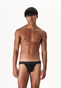String noir en tissu lisse, avec une taille large. Le vêtement épouse la silhouette, mettant en valeur le physique du mannequin.