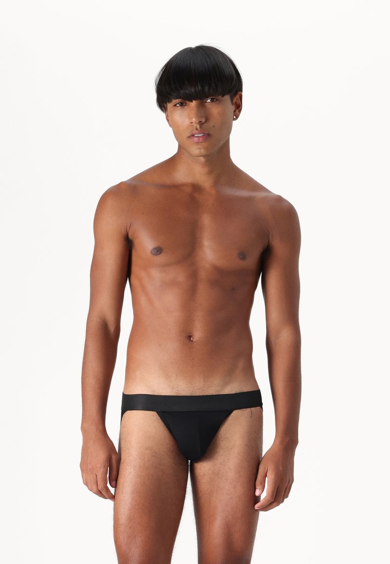 String noir en tissu lisse, avec une taille large. Le vêtement épouse la silhouette, mettant en valeur le physique du mannequin.