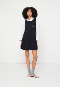 Mornarsko plava nabrana haljina s uskim korzetom i džepom s zlatnim logotipom. Ispod bijela košulja dugih rukava i svijetlosive čarape.