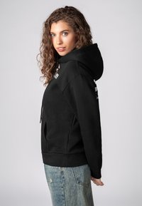 Schwarzer Kapuzenpullover aus einer Baumwollmischung, mit einer Fronttasche, gerippten Bündchen und einer lockeren Passform, kombiniert mit ausgewaschenen blauen Jeans.