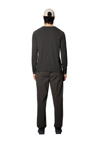 Mann trägt eine beige Baseballkappe, ein dunkles langärmliges Hemd, lockere dunkle Hose und schwarze Schuhe, steht mit dem Rücken zum Betrachter vor einem weißen Hintergrund.