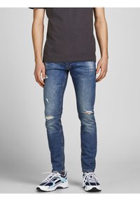 Hombre vestido con jeans ajustados azul desgastados, camiseta negra y zapatillas blanco-azul, de pie frente a un fondo liso.