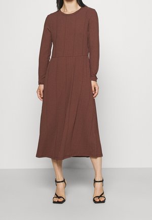 Woman wearing a long-sleeve, ribbed, brown midi dress with a round neckline and black ankle-strap high-heeled sandals.