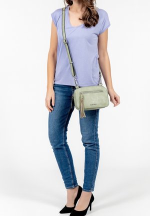 Lichtgroene suède crossbodytas met een rits, tassel-detail en verstelbare band, gecombineerd met een paars T-shirt en blauwe spijkerbroek.