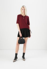 Cămașă polo bordeaux cu mâneci scurte și închidere cu nasturi, asociată cu o fustă plisată neagră. Ține în mână o geantă texturată neagră. Pantofi maro și negri.