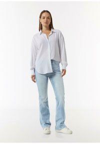 Witte lange mouwen overhemd met een ontspannen pasvorm, gecombineerd met lichtblauwe wijde jeans en witte sneakers, tegen een neutrale achtergrond.