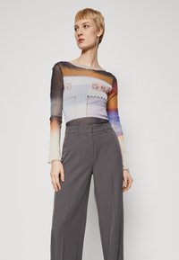 Maglietta a maniche lunghe in tessuto trasparente con un design astratto colorato; abbinata a pantaloni larghi grigi, che mettono in risalto una superficie liscia.