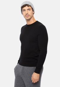 Schwarzer gestrickter Pullover mit Rundhalsausschnitt und langen Ärmeln, getragen über grauen, weichen Stoffhosen, kombiniert mit einer hellgrauen Mütze.