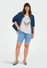Giacca in denim, t-shirt bianca con design grafico, pantaloni corti arrotolati di blu chiaro e tacchi turchesi. Attenzione agli strati casual e alla vestibilità rilassata.