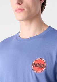 Light blue cotton t-shirt featuring a round red patch with "HUGO" text. Crew neck design with short sleeves and smooth texture.