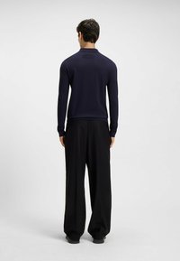 Mężczyzna stoi tyłem, na sobie ma granatową koszulę z długim rękawem oraz luźne czarne spodnie, w tle jasne, jednolite tło.