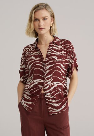 Kratka majica bordo barve s kratkimi rokavi in belimi črtami zebre, z ovratnikom, z gumbi spredaj in teksturirano tkanino. Nošena z ujemajočimi bordo hlačami.