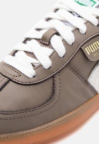 Krupan prikaz sivih Puma tenisica s bijelim vezicama, zlatnim Puma Paris logotipom i prozirnom smeđom gumom potplatom.