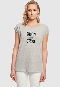 Grå t-skjorte med kort erme og løs passform, med svart tekst som leser "BLOOM and GROW" i sentrum. Enkelt stoffmønster.
