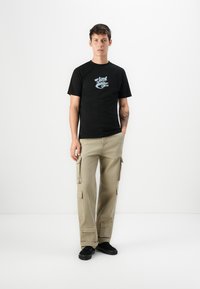 Zwarte t-shirt met lichtblauwe grafische print, gecombineerd met beige cargobroek met meerdere zakken en een relaxed fit. Zwart schoeisel maakt de outfit compleet.